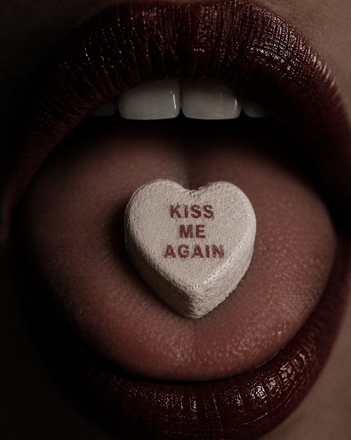 a tongue sticks out of dark red lips with a candy heart with the words kiss me again attracting a better kisser